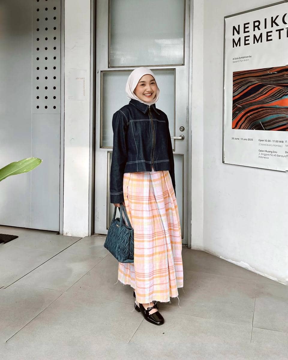Seorang wanita berhijab mengenakan jaket denim biru tua dan rok panjang motif kotak pastel sambil membawa tas biru di depan pintu kaca.