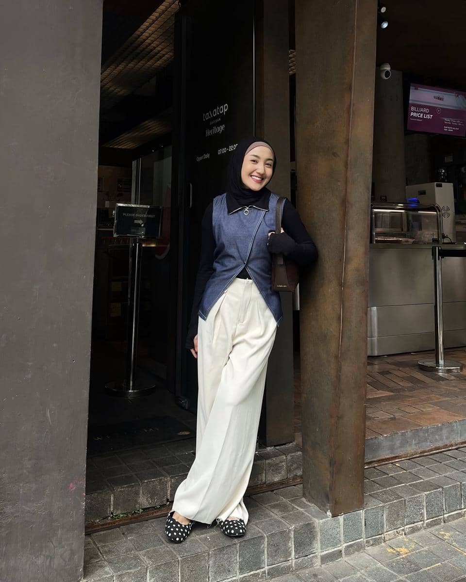 Seorang wanita berhijab mengenakan atasan denim tanpa lengan, inner hitam, celana panjang putih, dan sepatu hitam bermotif polkadot.