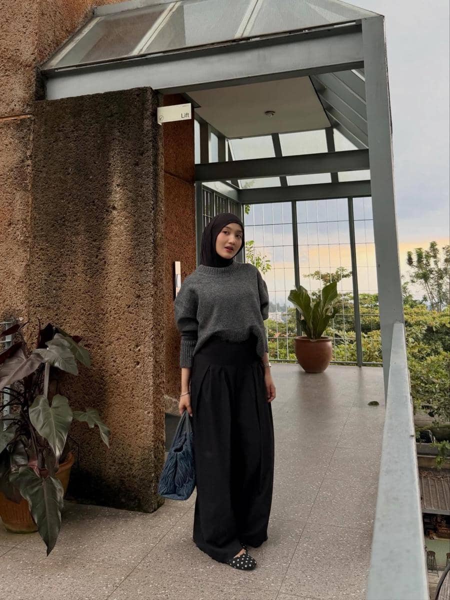 Seorang wanita berhijab mengenakan sweater abu-abu dan celana hitam longgar sambil memegang tas biru di area balkon dengan pemandangan hijau.