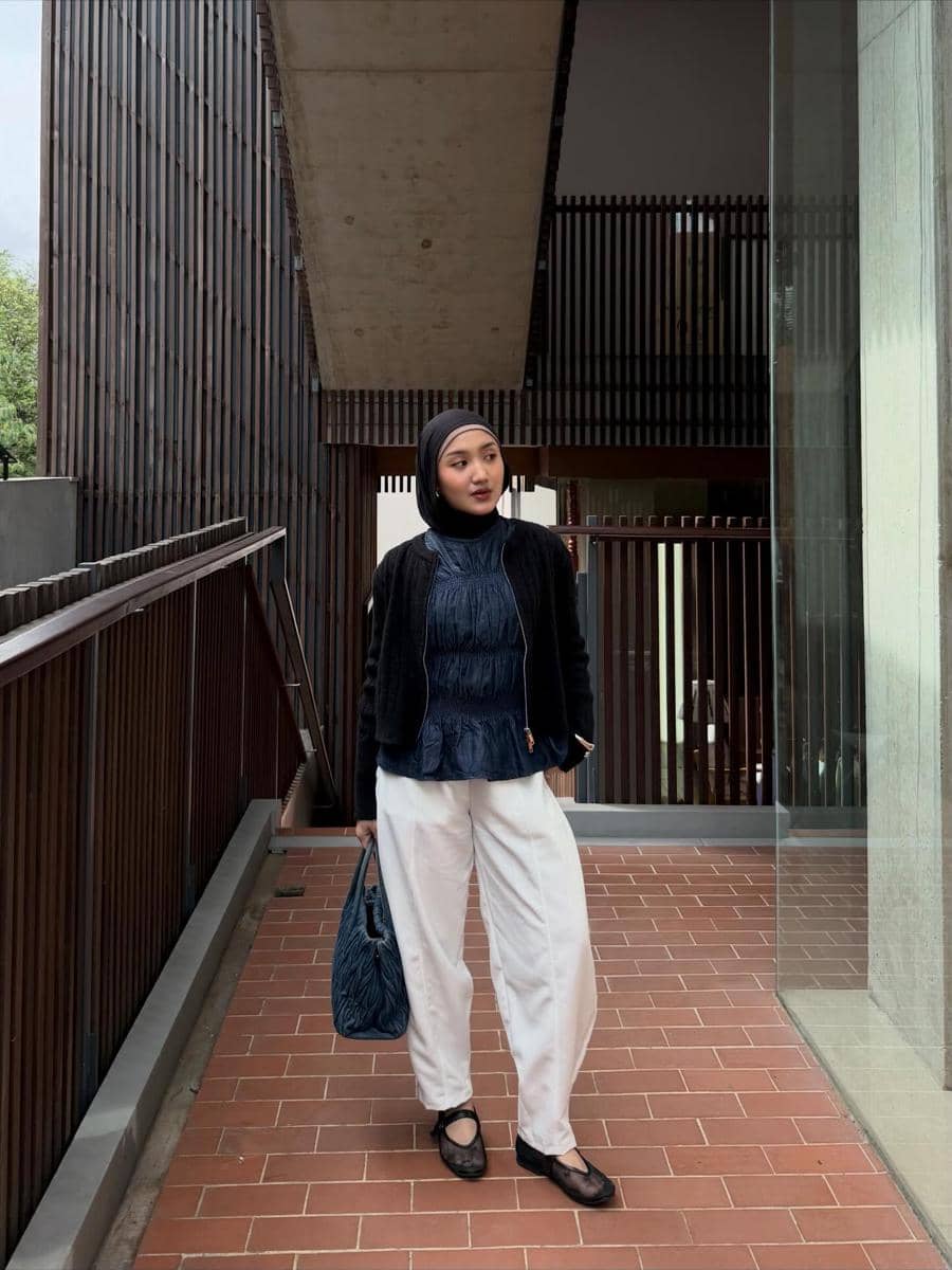 Seorang wanita berhijab mengenakan atasan denim biru tua, jaket hitam, celana putih longgar, dan membawa tas biru di area bangunan modern.