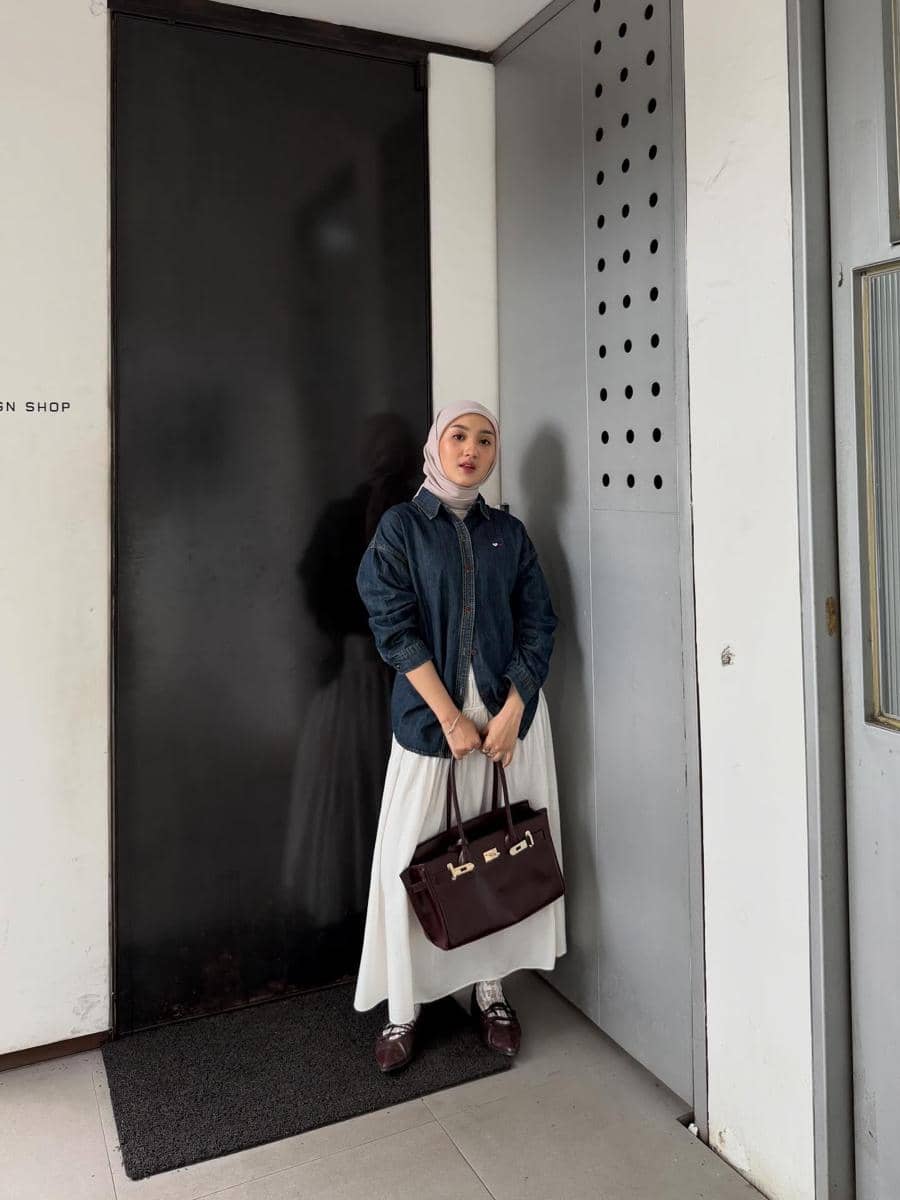 Seorang wanita berhijab mengenakan jaket denim biru, rok putih panjang, dan membawa tas tangan cokelat tua di depan pintu hitam.