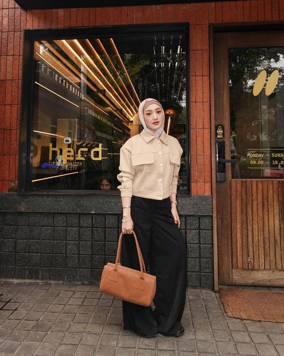 Seorang wanita berhijab mengenakan kemeja krem dan celana hitam lebar sambil membawa tas cokelat di depan kafe bergaya modern.