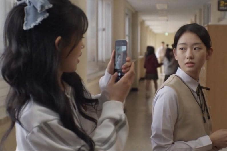 Dua siswi berbicara di koridor sekolah, salah satunya memegang ponsel untuk mengambil foto dalam adegan drama Agent Kim Reactivated.