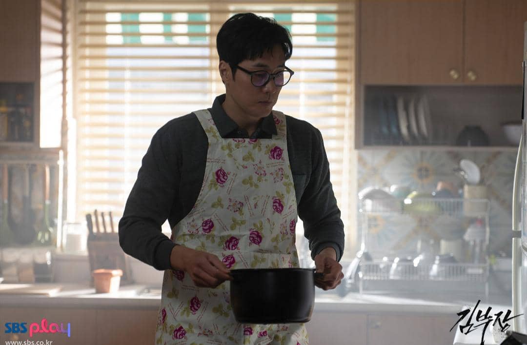 Seorang pria mengenakan celemek bermotif bunga sedang memegang panci di dapur dengan pencahayaan lembut dari jendela.