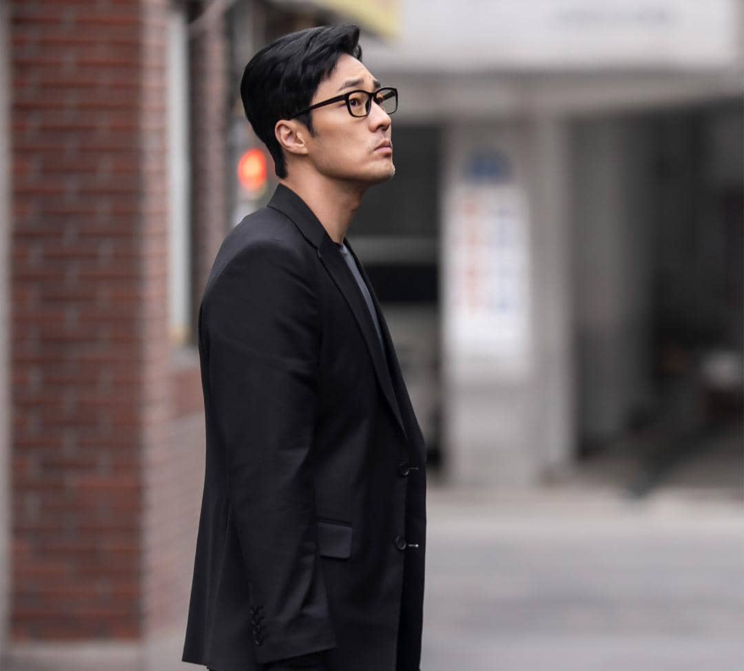 Seorang pria mengenakan setelan jas hitam dan kacamata berdiri di jalan sempit, dalam cuplikan drama Agent Kim Reactivated.