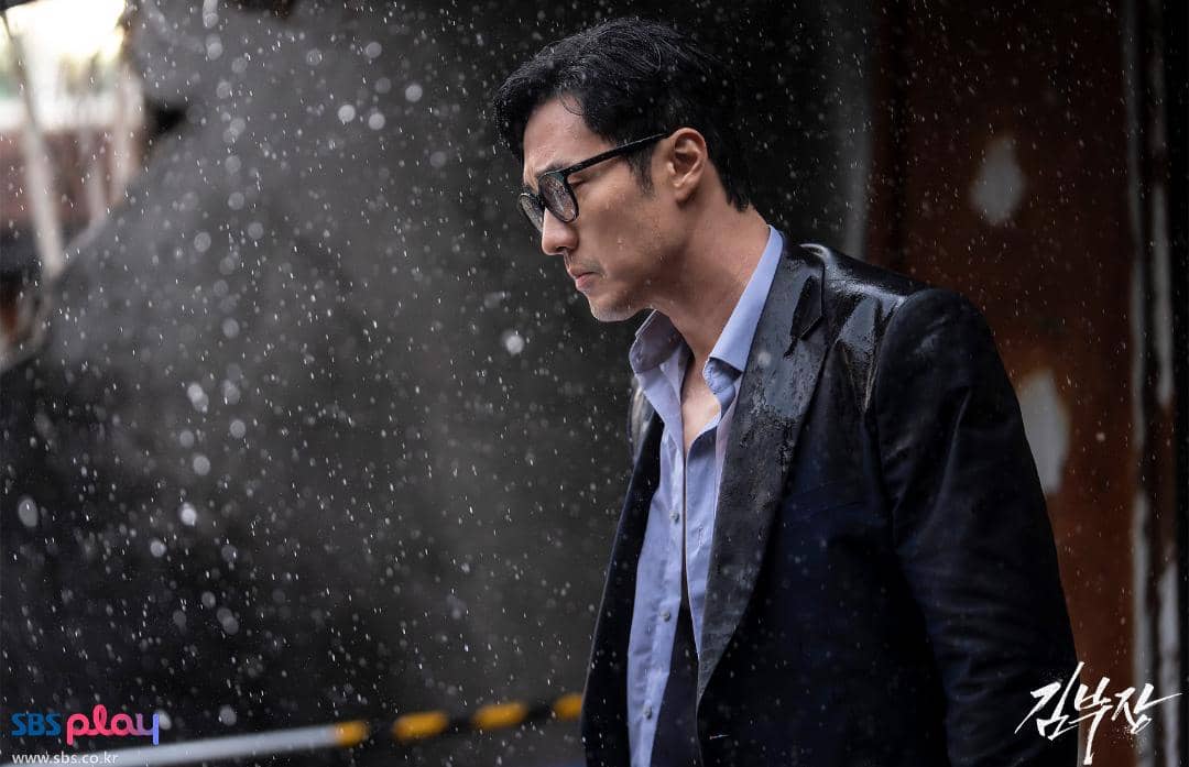 Seorang pria berjas hitam dan berkacamata berdiri di bawah hujan deras dengan ekspresi serius dalam cuplikan drama Agent Kim Reactivated.