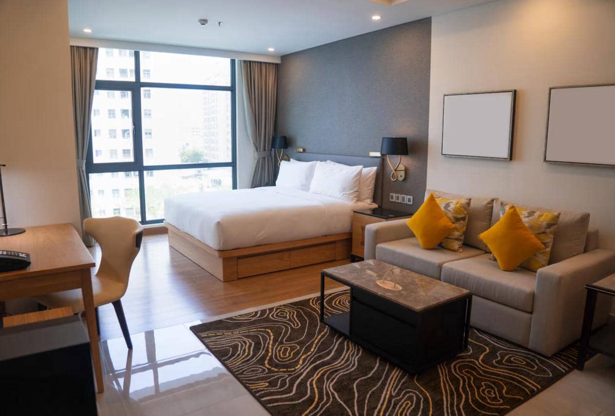 Kamar guest house modern dengan tempat tidur besar, sofa abu-abu beraksen bantal kuning, meja kerja kayu, dan jendela besar berpemandangan kota.