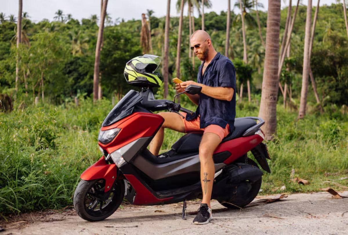 Seorang traveler duduk di atas motor skuter merah sambil memegang ponsel di area tropis dengan latar pepohonan kelapa.