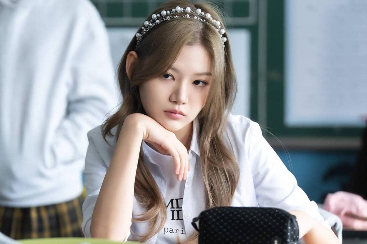 Seorang perempuan berambut panjang mengenakan bando mutiara duduk di kelas dengan ekspresi serius dalam cuplikan drama Agent Kim Reactivated.
