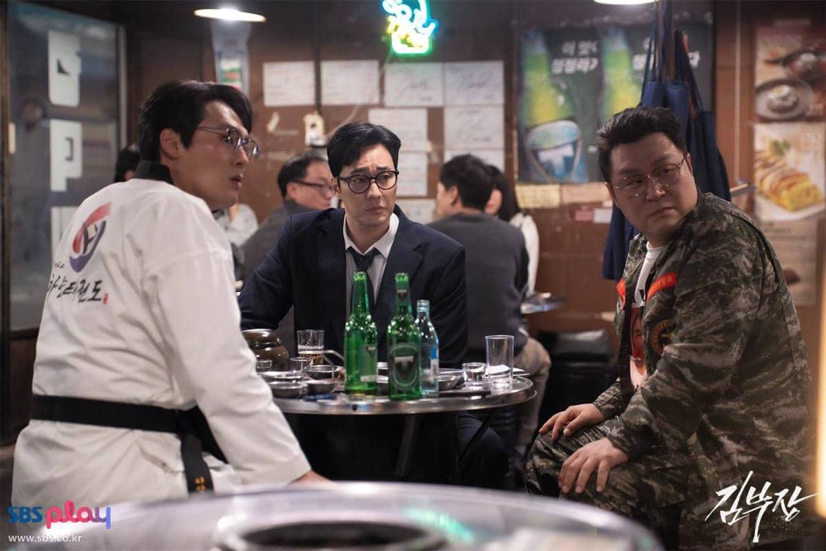 Tiga pria duduk di meja restoran dengan suasana malam, mengenakan pakaian berbeda termasuk jas, seragam taekwondo, dan jaket loreng.