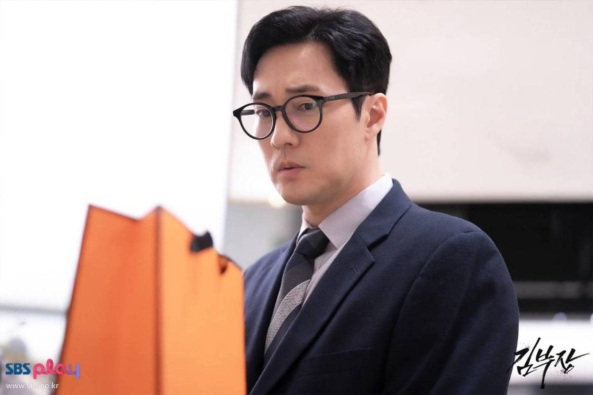 Seorang pria mengenakan setelan jas biru tua dan kacamata hitam bulat sedang memandang tas oranye di depannya dalam adegan drama Agent Kim Reactivated.