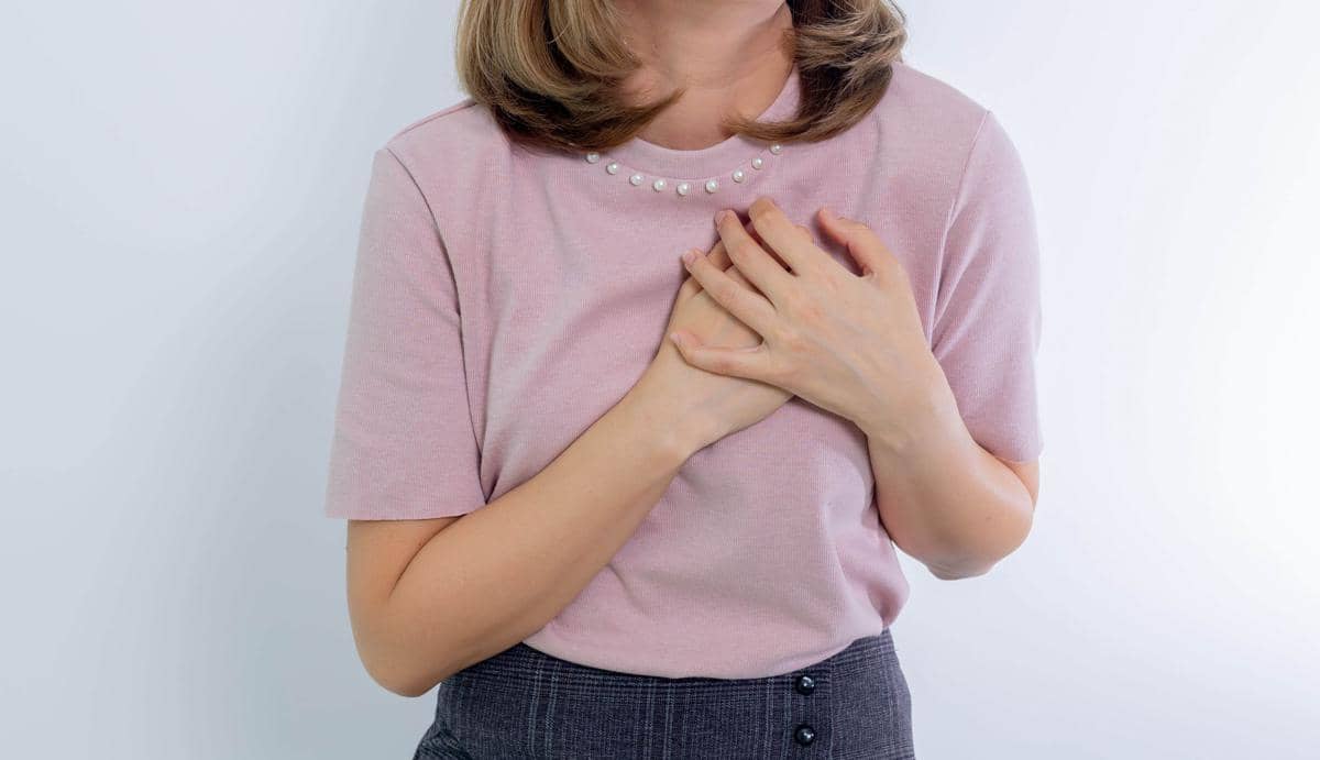 Seorang wanita mengenakan kaus ungu muda memegang dadanya dengan kedua tangan, menggambarkan rasa tidak nyaman pada area payudara.