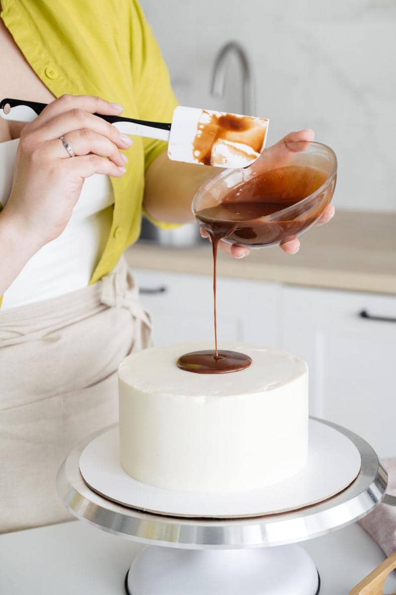 Seseorang menuangkan ganache cokelat di atas kue putih menggunakan spatula di dapur modern dengan meja putar kue.
