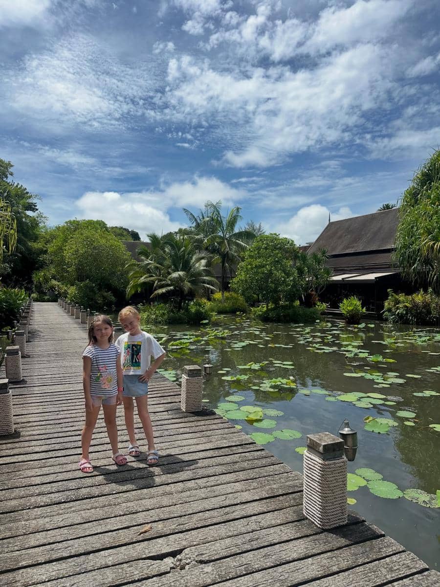 Dua anak berdiri di jembatan kayu di atas kolam berisi daun teratai dengan latar pepohonan hijau dan langit biru cerah di Phuket.