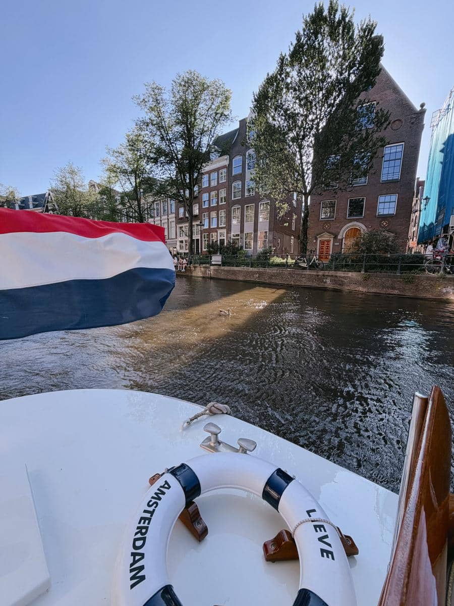 Pemandangan kanal di Amsterdam dengan rumah-rumah khas Belanda, bendera Belanda di perahu, dan pelampung bertuliskan Amsterdam.