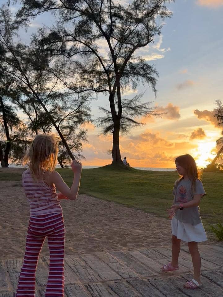 Dua anak bermain di tepi pantai saat matahari terbenam di Phuket dengan latar pepohonan dan langit berwarna jingga keemasan.