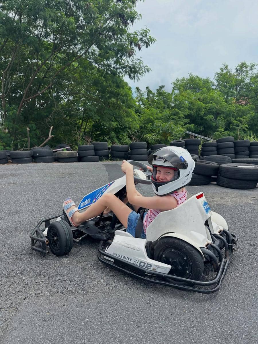 Seorang anak mengenakan helm sedang mengendarai gokart putih di lintasan luar ruangan dengan latar ban hitam dan pepohonan hijau.