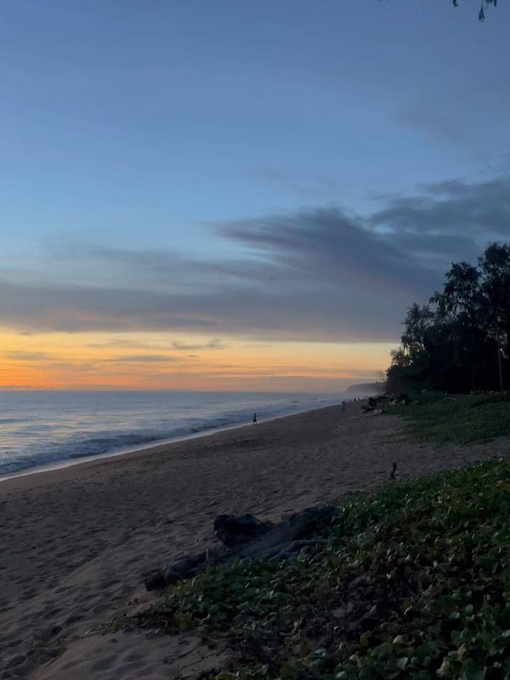 Pemandangan pantai di Phuket saat matahari terbenam dengan langit berwarna jingga dan biru serta garis pantai yang tenang.