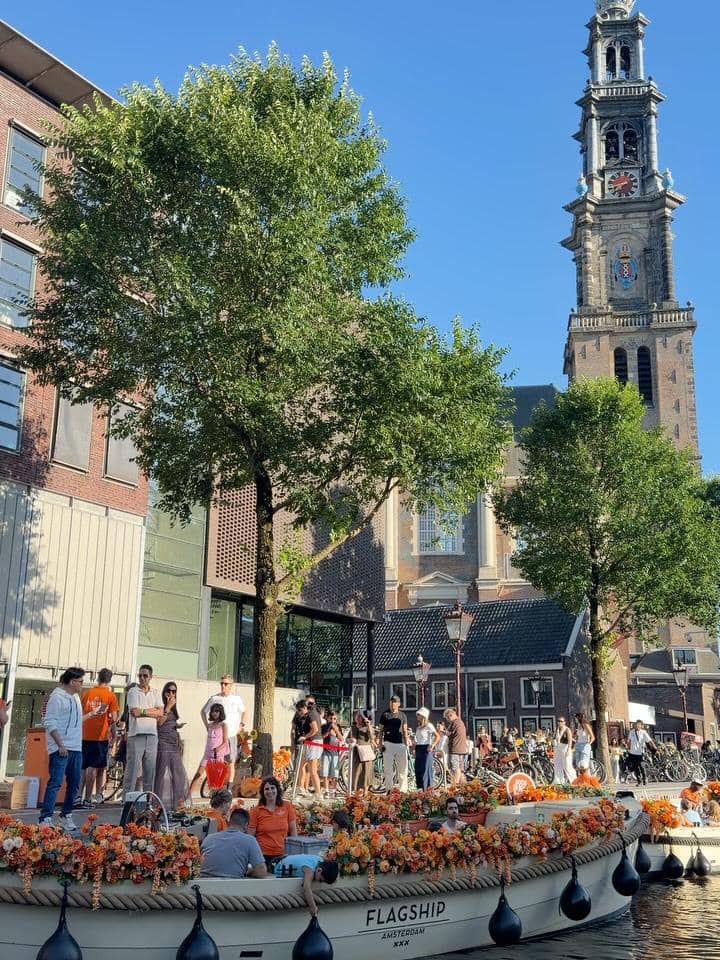 Orang-orang menikmati wisata perahu di kanal Amsterdam dengan dekorasi bunga oranye, berlatar bangunan bersejarah dan menara gereja.