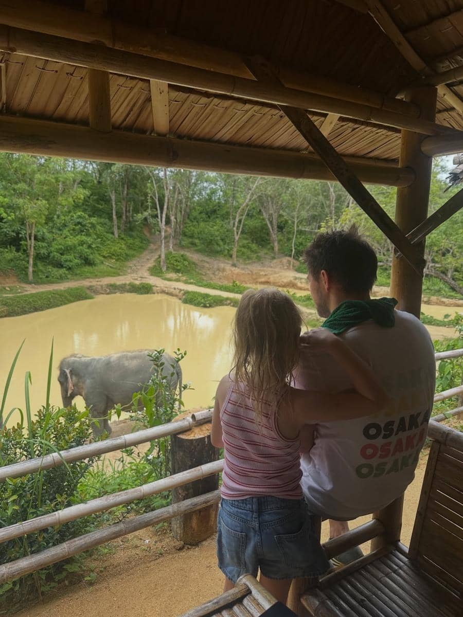 Seorang pria dan anak kecil berdiri di teras bambu menghadap kolam berlumpur dengan seekor gajah di tengah area hijau di Phuket.