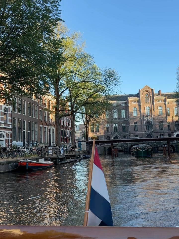 Pemandangan kanal di Belanda dengan bendera Belanda di perahu, dikelilingi bangunan klasik dan pepohonan di bawah langit cerah.