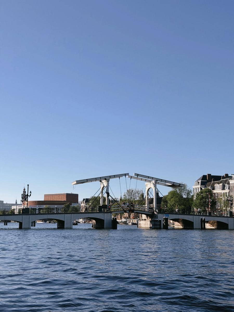 Jembatan putih bergaya klasik di atas kanal dengan air tenang dan bangunan berarsitektur Eropa di bawah langit biru cerah.