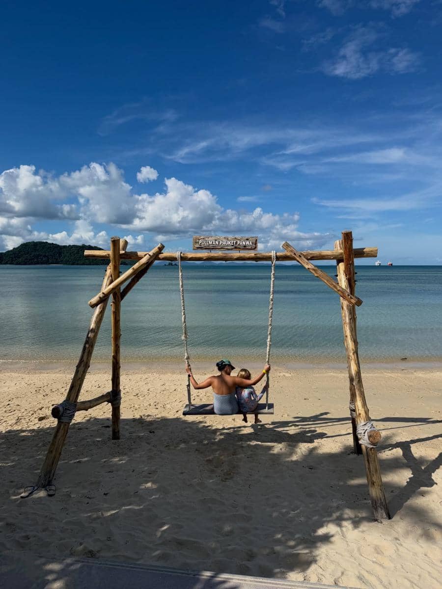 Seorang wanita dan anak kecil duduk di ayunan kayu di tepi pantai berpasir dengan latar laut biru dan langit cerah di Phuket.