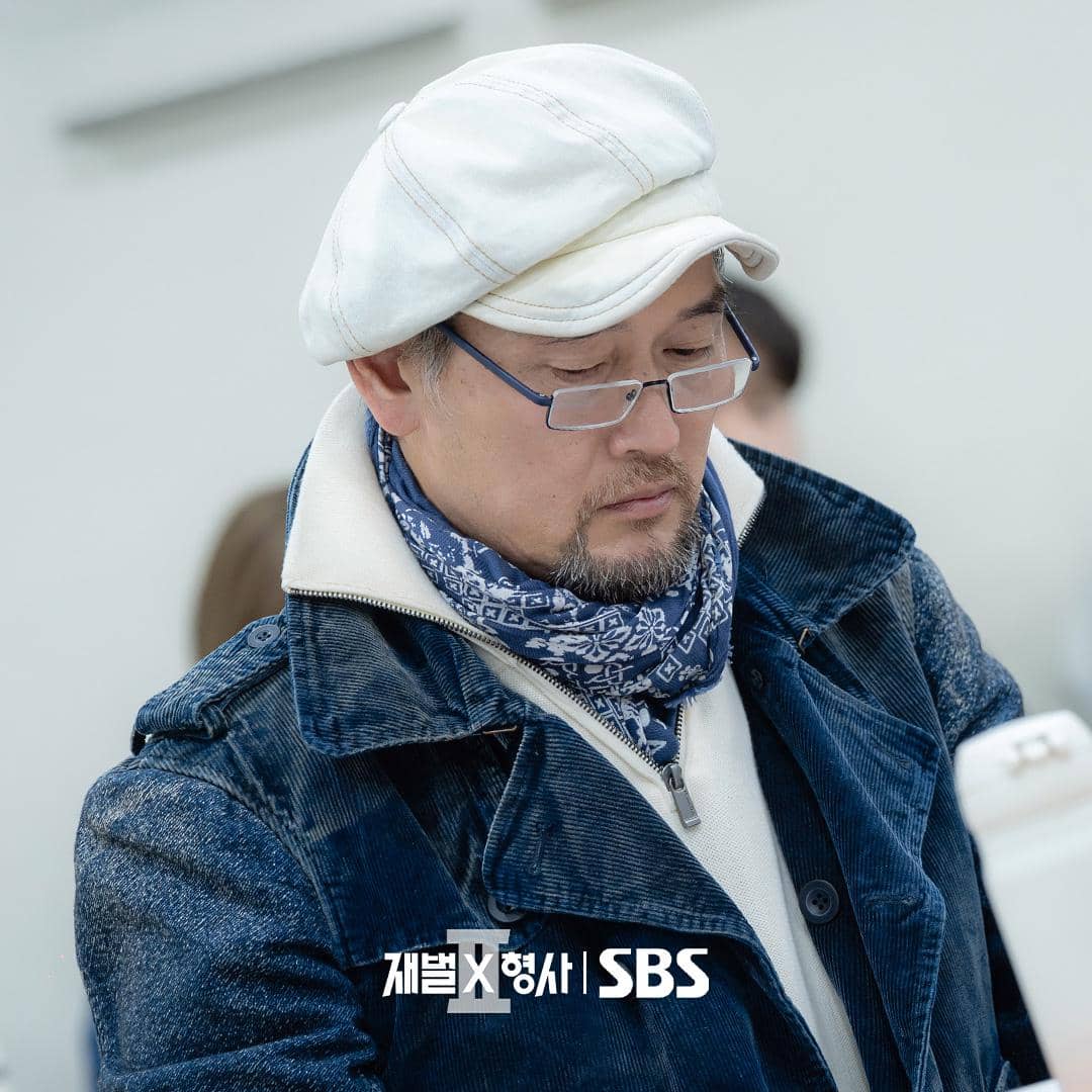 Seorang pria mengenakan topi putih, kacamata, dan jaket biru sedang membaca naskah dalam sesi pembacaan drakor Flex X Cop 2.