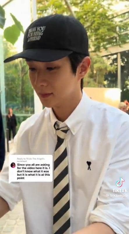 Seorang pria mengenakan topi hitam bertulisan putih, kemeja putih, dan dasi hitam putih sedang berada di luar ruangan dengan latar pepohonan.