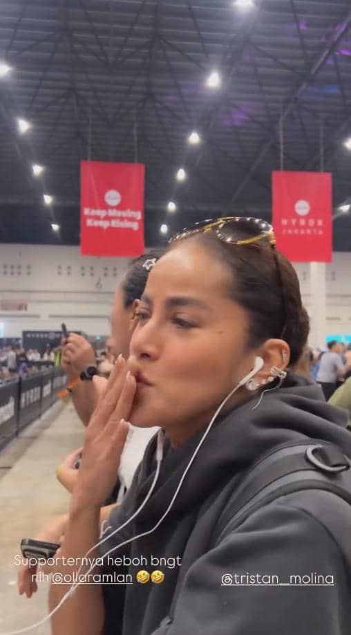 Seorang wanita mengenakan hoodie abu-abu dan kacamata di kepala tampak memberi dukungan di acara HYROX Jakarta 2026 di dalam gedung besar.