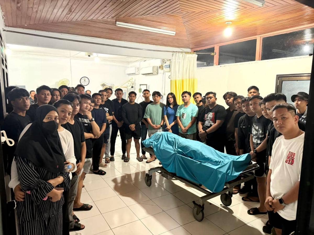 Keluarga dan teman-teman berdiri mengelilingi jenazah yang ditutupi kain biru di ruang rumah sakit RSUD Bolmut pada malam hari.