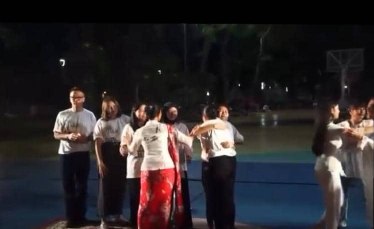 Sekelompok orang mengenakan pakaian putih dan hitam sedang berpelukan serta berdoa bersama di area terbuka pada malam hari.