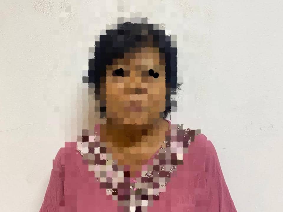 Perempuan berusia lanjut mengenakan baju merah muda dengan wajah disamarkan, berdiri di depan dinding berwarna putih.