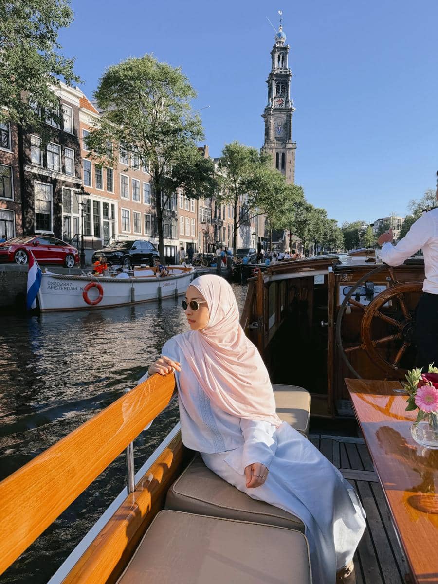Seorang perempuan berhijab duduk santai di atas perahu kayu di kanal Amsterdam dengan latar menara gereja dan bangunan klasik Belanda.