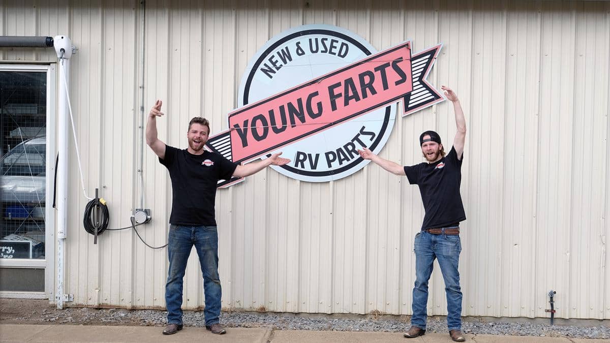 Dua pria berdiri di depan dinding bergambar logo besar bertuliskan Young Farts RV Parts dengan ekspresi gembira dan tangan terangkat.
