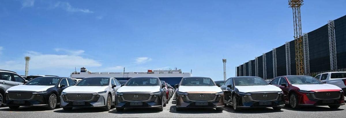 Deretan mobil Mazda 6e baru berbagai warna terparkir rapi di area pelabuhan dengan latar kapal dan langit cerah.