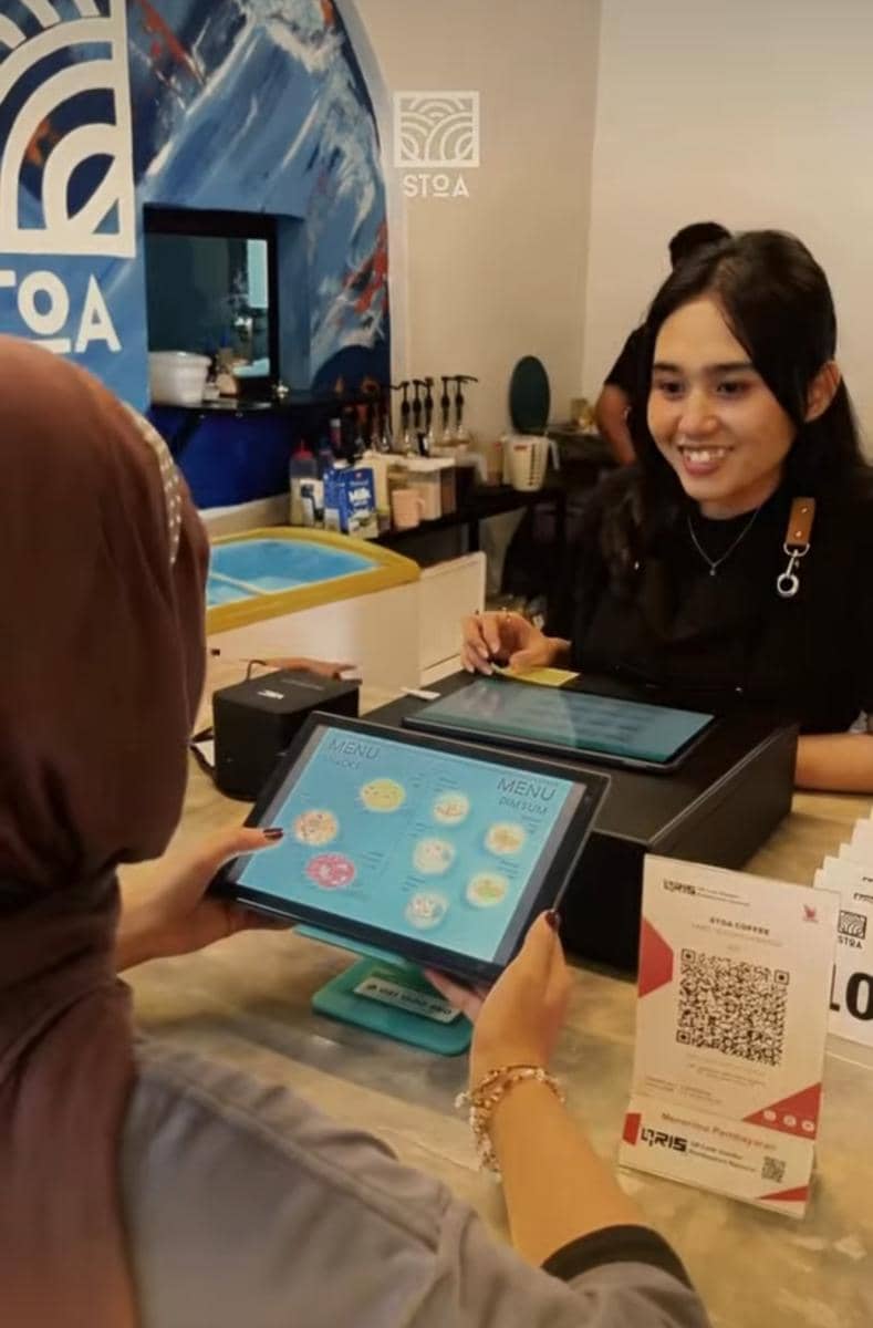 Barista tersenyum melayani pelanggan yang memesan minuman menggunakan tablet di konter Stoa Coffee Books dengan latar interior biru putih.