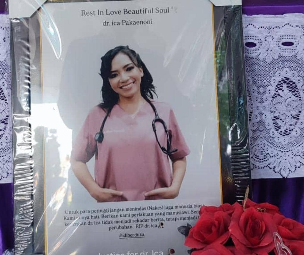 Potret dr. Icha dengan seragam medis berwarna merah muda dan stetoskop di leher, dipajang di rumah duka dengan hiasan bunga merah di depannya.