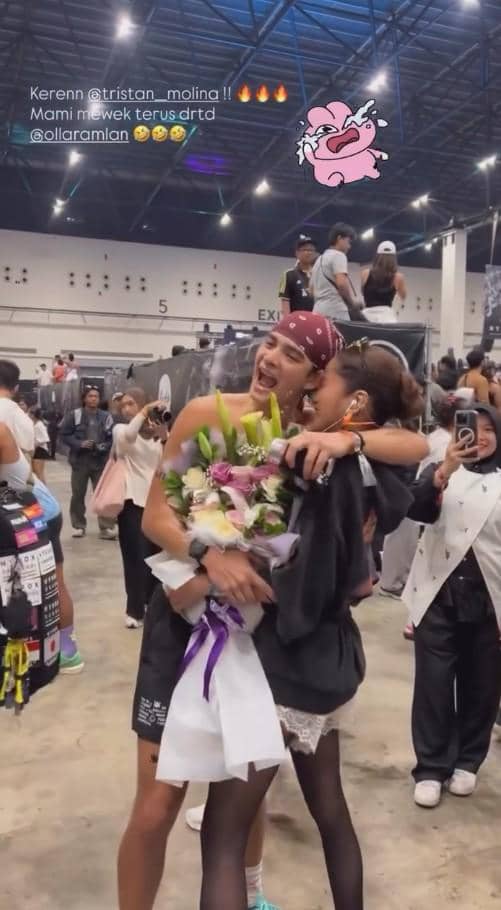Seorang pria memeluk wanita sambil memegang buket bunga di acara olahraga dalam ruangan dengan banyak penonton di sekitarnya.