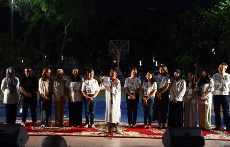 Sekelompok orang berdiri di atas panggung luar ruangan saat malam hari, mengikuti doa bersama menjelang putusan terkait Nadiem Makarim.