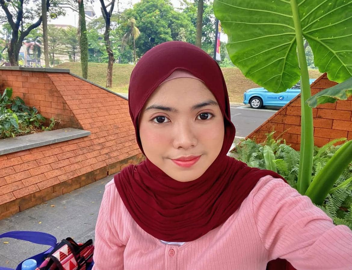 Perempuan berhijab merah marun mengenakan atasan merah muda berfoto selfie di area taman dengan latar pepohonan dan dinding bata oranye.