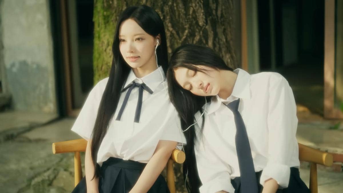 Dua gadis mengenakan seragam sekolah duduk berdampingan di luar ruangan, satu bersandar di bahu lainnya sambil berbagi earphone.