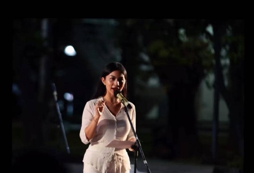 Seorang perempuan mengenakan pakaian putih berbicara di depan mikrofon sambil memegang kertas pada acara doa bersama di malam hari.