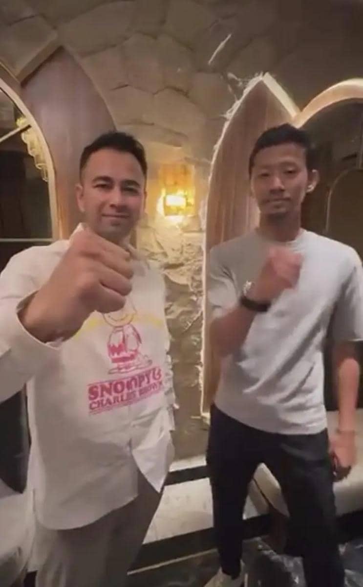 Dua pria berdiri berdampingan di dalam ruangan dengan dinding batu dan pencahayaan hangat, keduanya mengangkat tangan dengan gestur salam.