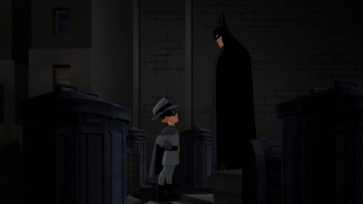 Batman berdiri di gang gelap berbicara dengan anak kecil berpakaian abu-abu dan bertopi dalam adegan animasi Batman: Caped Crusader Season 2.