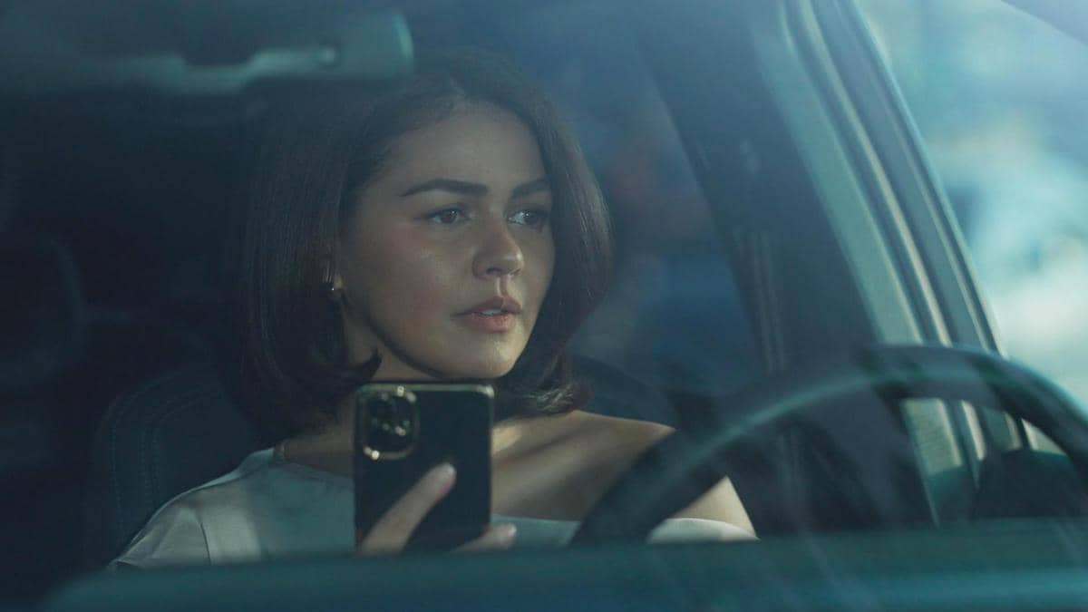 Seorang perempuan duduk di dalam mobil sambil memegang ponsel, terlihat serius menatap ke arah luar jendela di suasana redup.
