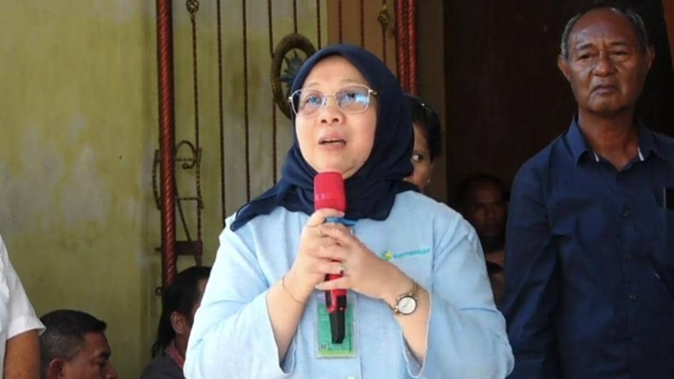 Seorang perempuan mengenakan kerudung biru dan kemeja biru muda memegang mikrofon merah saat berbicara di depan beberapa orang.