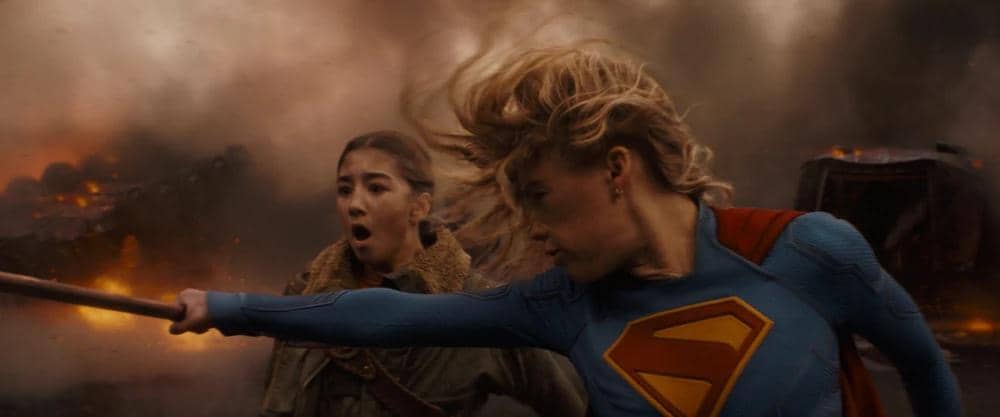 Supergirl mengenakan kostum biru dengan lambang S di dada sedang bertarung di tengah ledakan besar bersama seorang wanita di latar belakang.