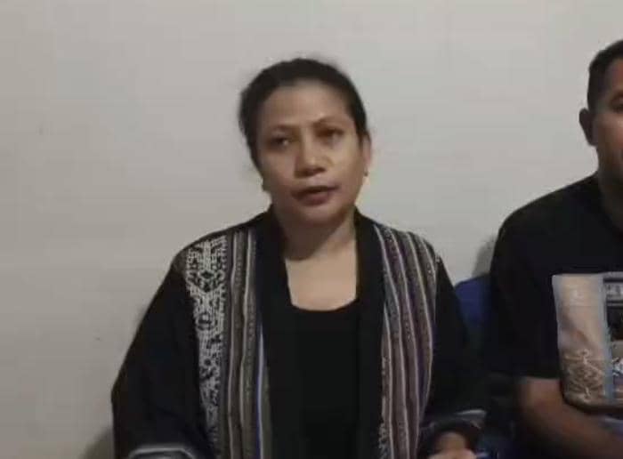 Seorang perempuan mengenakan pakaian hitam bermotif duduk di samping seorang pria di ruangan dengan dinding polos.
