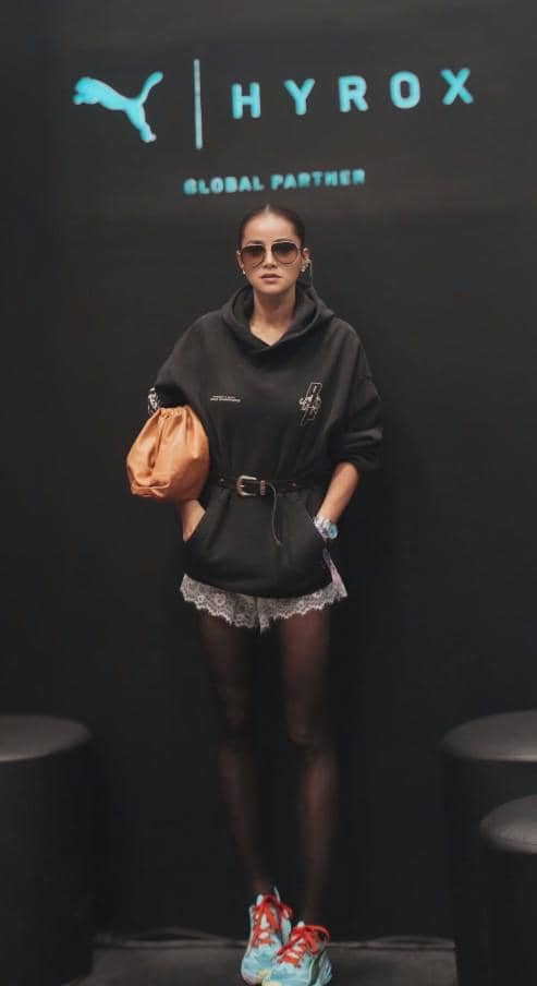 Seorang wanita bergaya kasual mengenakan hoodie hitam, celana pendek renda, dan sepatu olahraga berpose di depan latar bertuliskan HYROX.